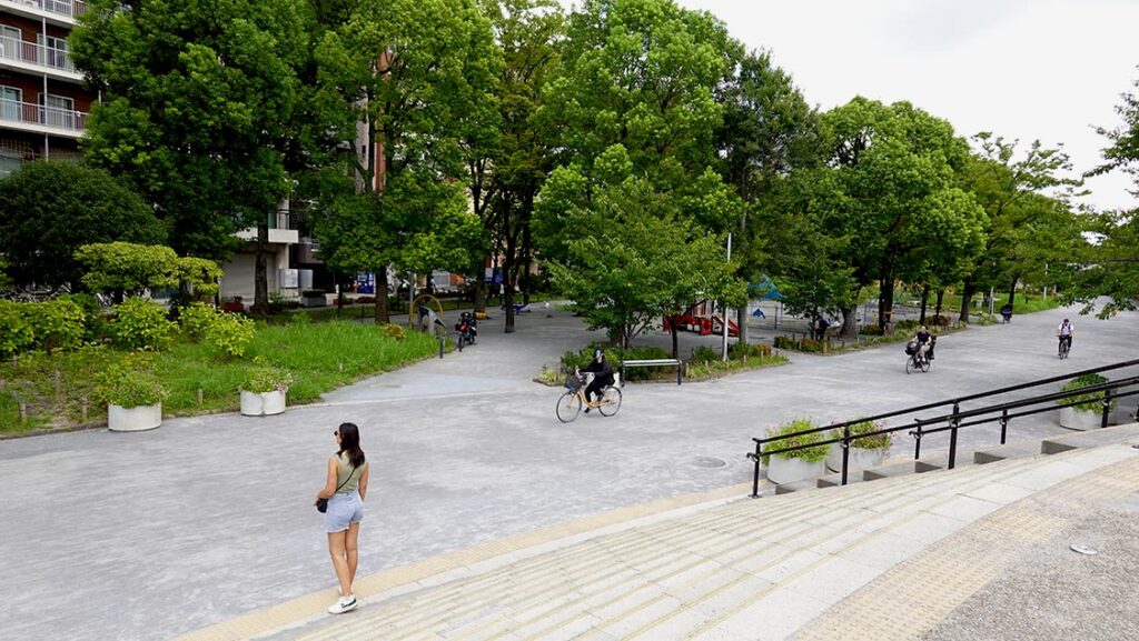 Noelia en el Parque Sumida con árboles y un ciclista pasando por el camino junto al río.
