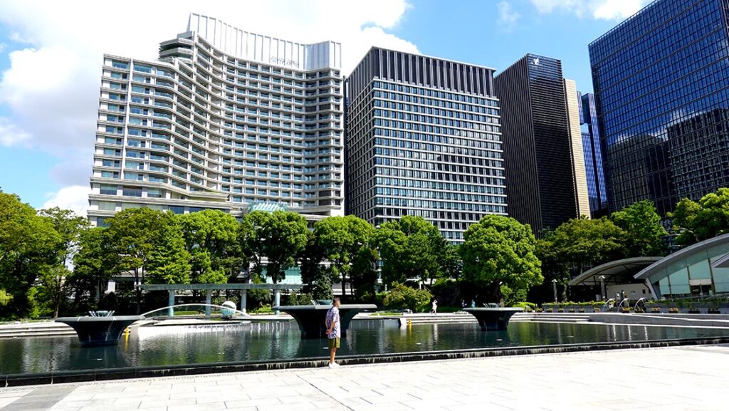 Joan sentado en el Parque de la Fuente de Wadakura en Tokio con los canales de agua y torres de cristal al fondo.