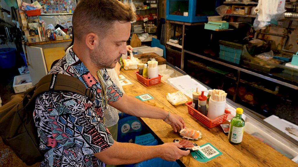 Joan comiendo piezas de sushi fresco de pie en la barra de un puesto tradicional del mercado de Tsukiji.
