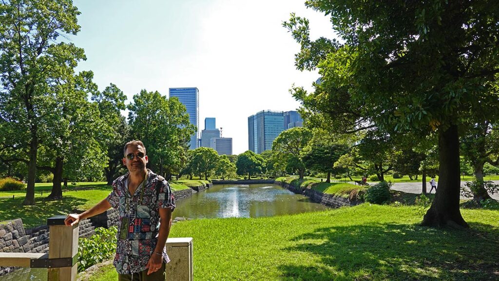 Joan cruzando un puente de piedra sobre el estanque de los Jardines de Hamarikyu con rascacielos de Tokio de fondo.